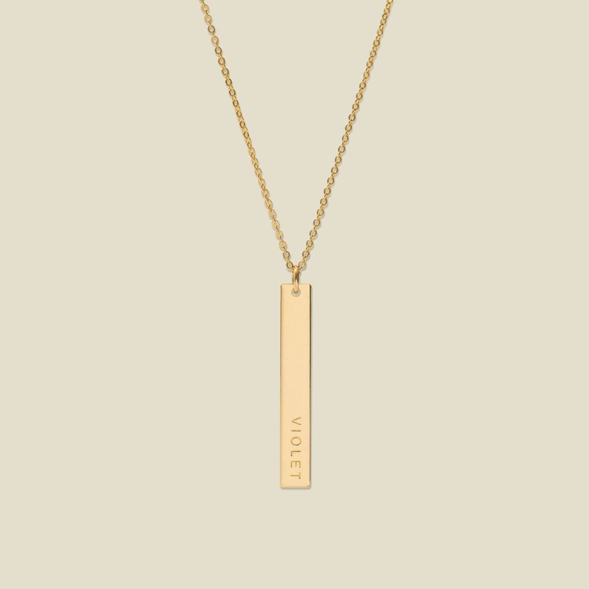 A gold pendant necklace with a vertical bar engraved with the name 'VIOLET' on a light beige background.