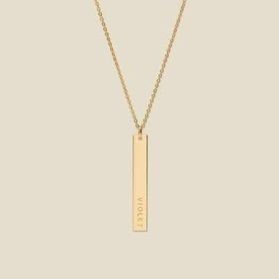 A gold pendant necklace with a vertical bar engraved with the name 'VIOLET' on a light beige background.