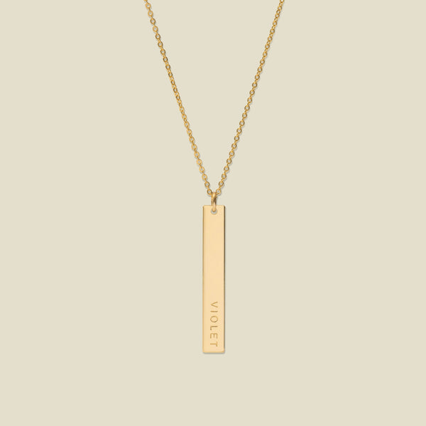 A gold pendant necklace with a vertical bar engraved with the name 'VIOLET' on a light beige background.