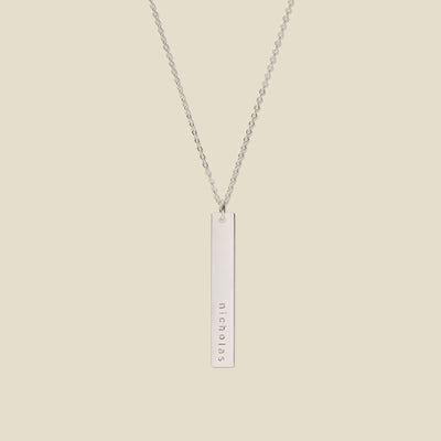 A silver pendant necklace with a vertical bar engraved with the name 'nicholas', shown on a solid light beige background.