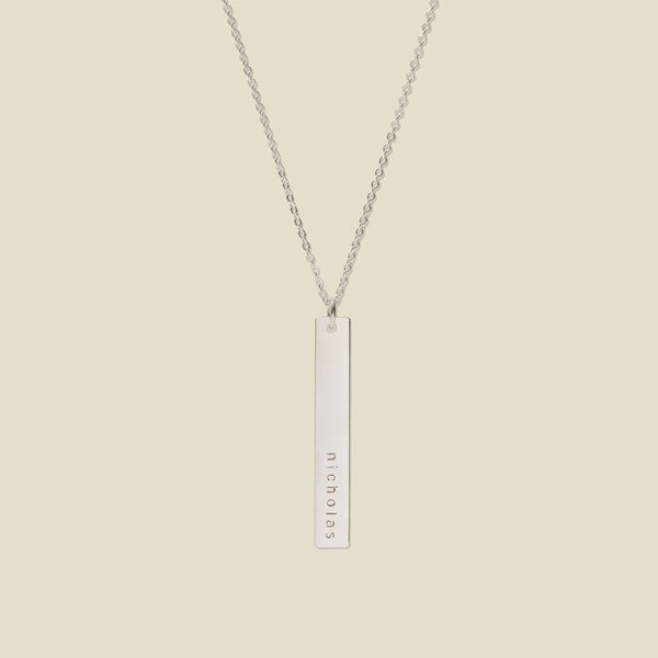 A silver pendant necklace with a vertical bar engraved with the name 'nicholas', shown on a solid light beige background.