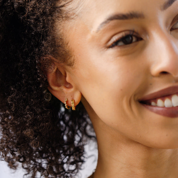 A close-up of a person's ear wearing two gold hoop earrings, one with clear round gemstones in a curving pattern and the other with the word 'VIRGO' in raised letters across the front.