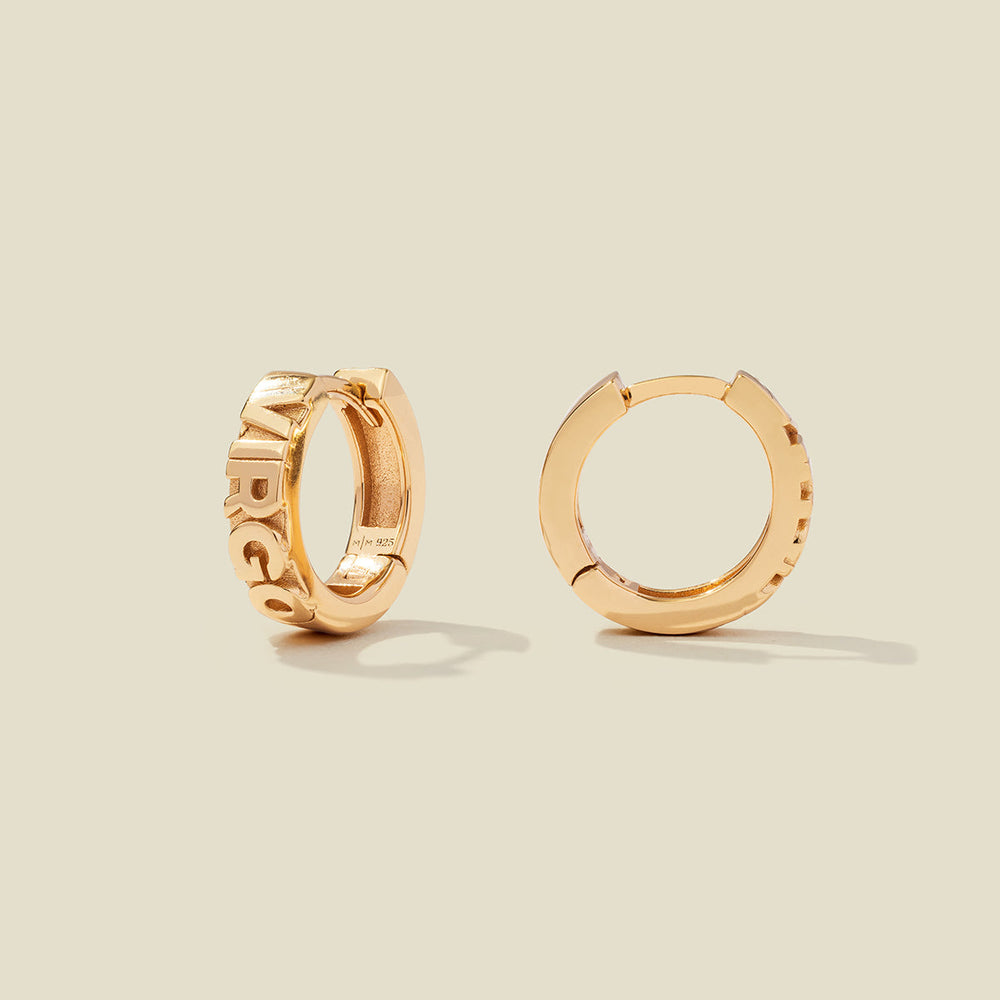 A pair of gold huggie hoop earrings on a light beige background. One earring is shown from the side, displaying a polished outer surface, while the other is shown from the front, showcasing the word 'VIRGO' in raised letters.