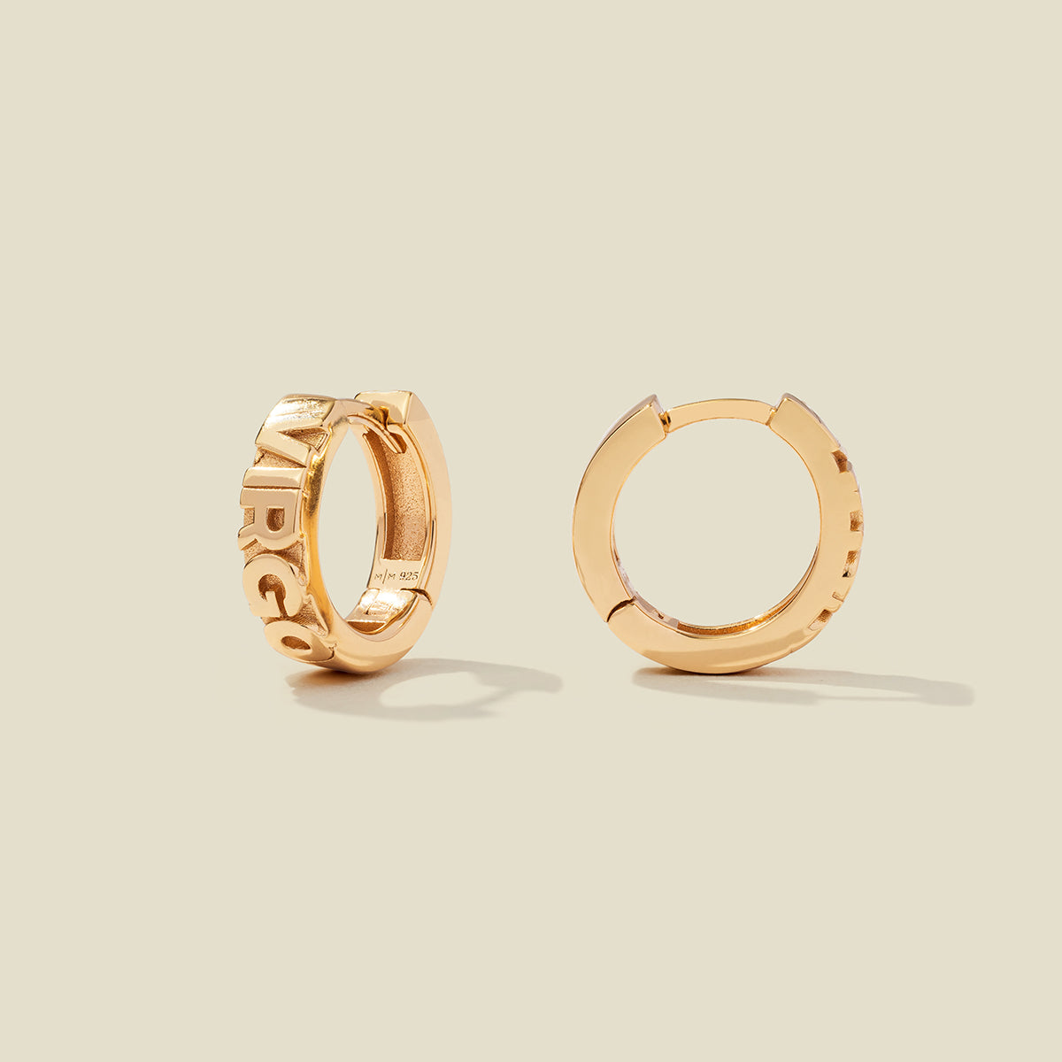 A pair of gold huggie hoop earrings on a light beige background. One earring is shown from the side, displaying a polished outer surface, while the other is shown from the front, showcasing the word 'VIRGO' in raised letters.