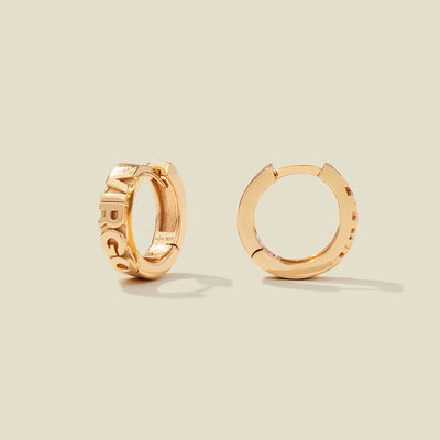 A pair of gold huggie hoop earrings on a light beige background. One earring is shown from the side, displaying a polished outer surface, while the other is shown from the front, showcasing the word 'VIRGO' in raised letters.