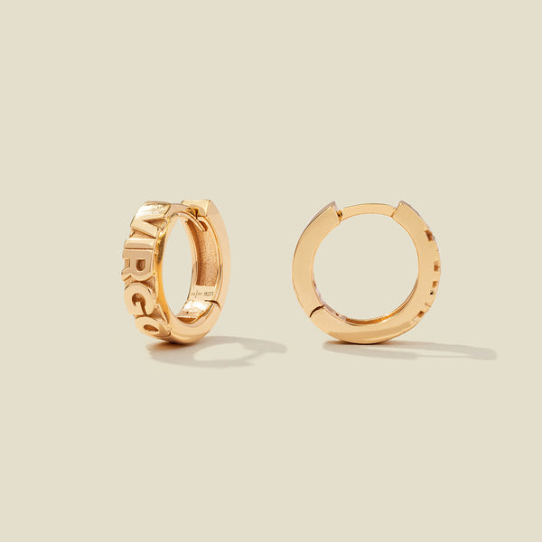 A pair of gold huggie hoop earrings on a light beige background. One earring is shown from the side, displaying a polished outer surface, while the other is shown from the front, showcasing the word 'VIRGO' in raised letters.