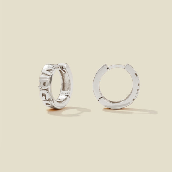 A pair of silver huggie hoop earrings on a light beige background. One earring is shown from the side, displaying a polished outer surface, while the other is shown from the front, showcasing the word 'VIRGO' in raised letters.