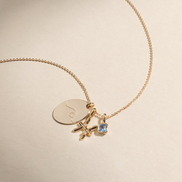 A gold delicate chain necklace with an oval pendant engraved with a wave design, a pisces symbol pendant with clear accent stones, and a light blue square gemstone charm.