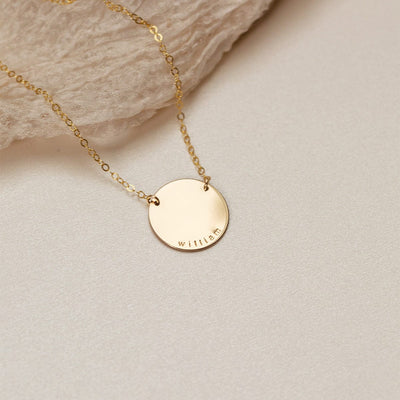 A gold chain necklace with a flat round disc charm engraved with the word 'william', shown on a textured light beige background with a small piece of light brown crinkled fabric.