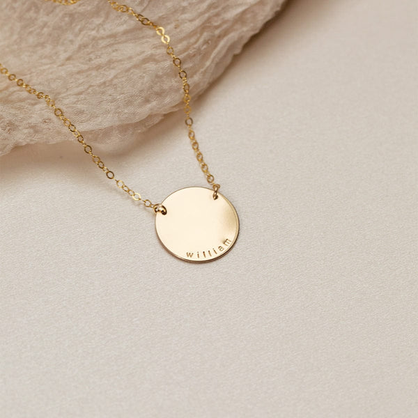 A gold chain necklace with a flat round disc charm engraved with the word 'william', shown on a textured light beige background with a small piece of light brown crinkled fabric.