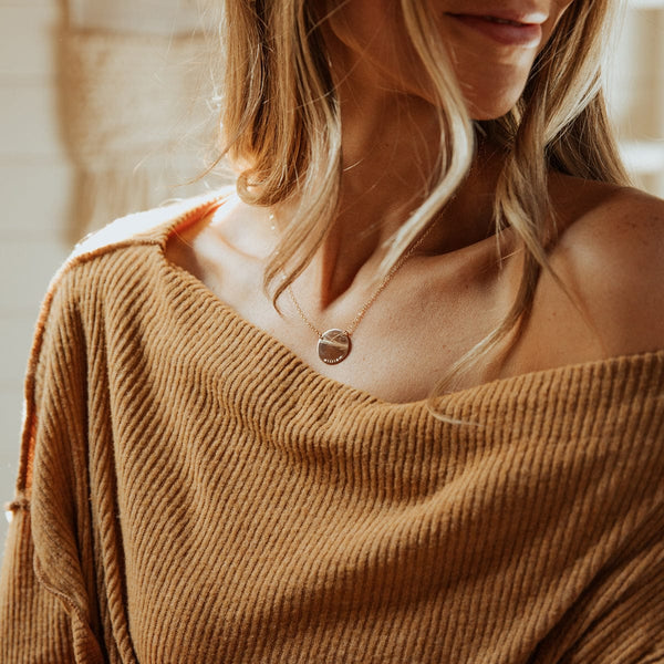 A woman in a brown off the shoulder ribbed knit top wearing a gold pendant chain with a flat round disc in the center.