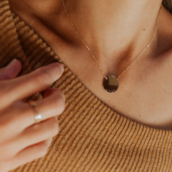 A person in a brown ribbed knit top wearing a delicate gold chain necklace with a flat round engraved disc connected in two spots to the chain.