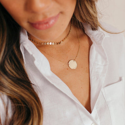 A person wearing a white button down shirt styled with a gold choker necklace made up of multiple flat circular discs and a longer gold chain necklace with an engraved flat circle.