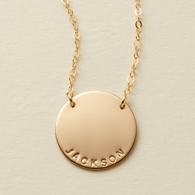 A gold personalized necklace featuring a flat round disc pendant engraved with the name 'JACKSON' at the bottom, suspended from a delicate chain in two spots against a light beige textured background.