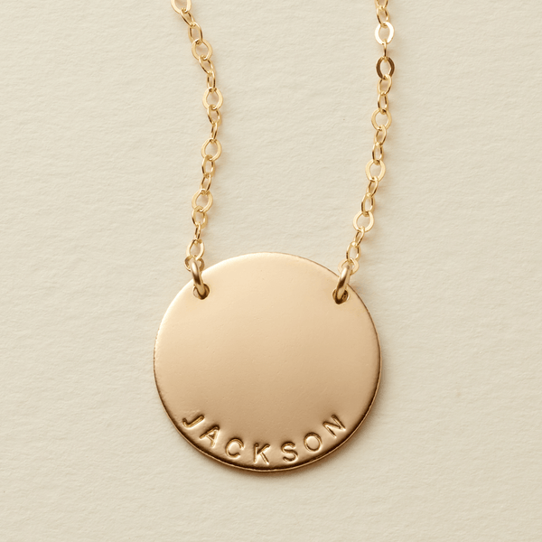 A gold personalized necklace featuring a flat round disc pendant engraved with the name 'JACKSON' at the bottom, suspended from a delicate chain in two spots against a light beige textured background.