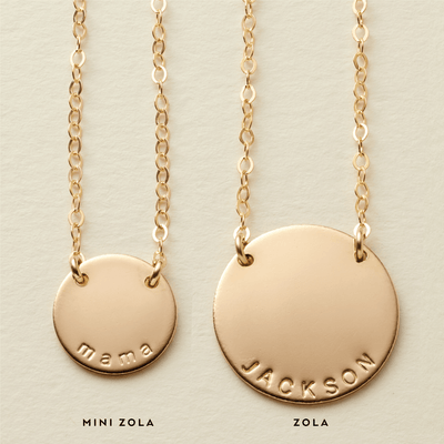 Two gold pendant necklaces, each with a different size of engraved round flat disc attached in two places to the chain. The smaller disc is engraved with the word 'mama' and has the title 'MINI ZOLA' below it, while the larger disc has the name 'JACKSON' engraved and has the word 'ZOLA' below it.