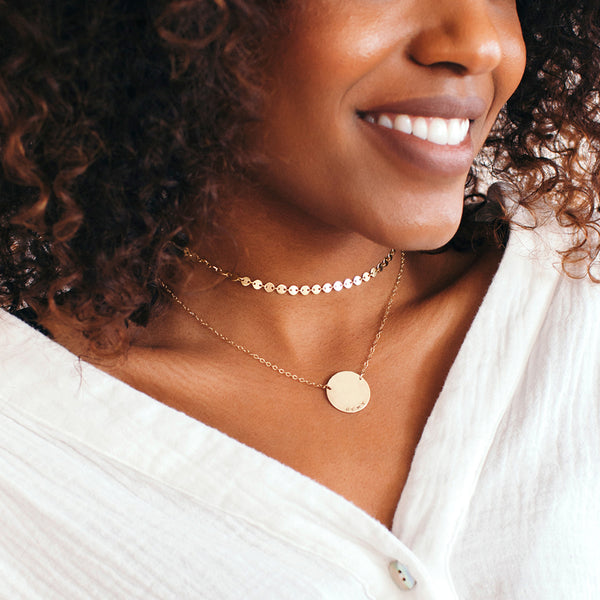 A close-up lifestyle shot of a smiling woman wearing two delicate gold necklaces: a short choker with small round discs and a longer chain with a flat circular disc pendant.